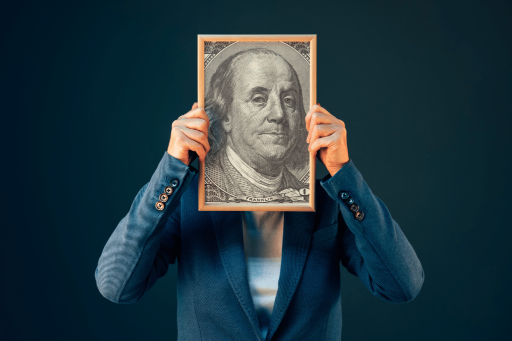 Person in blue blazer holding framed picture of Benjamin Franklin in front of her face.